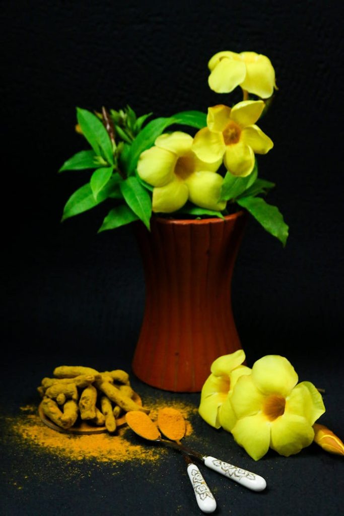 Yellow flowers in a vase with turmeric roots and powder on a dark background.